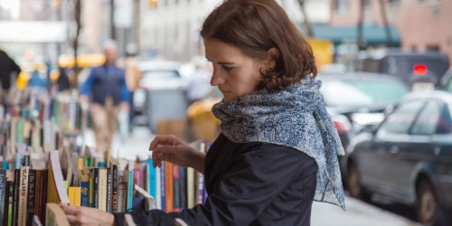 A woman browses books at an outdoor book stall on a city street, wearing a blue patterned scarf.