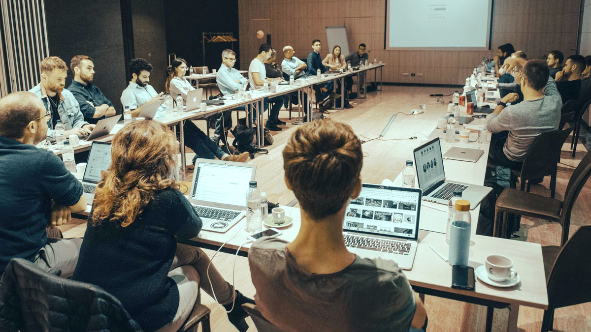 A large group meeting in a conference room with participants seated around a rectangular table, using laptops.