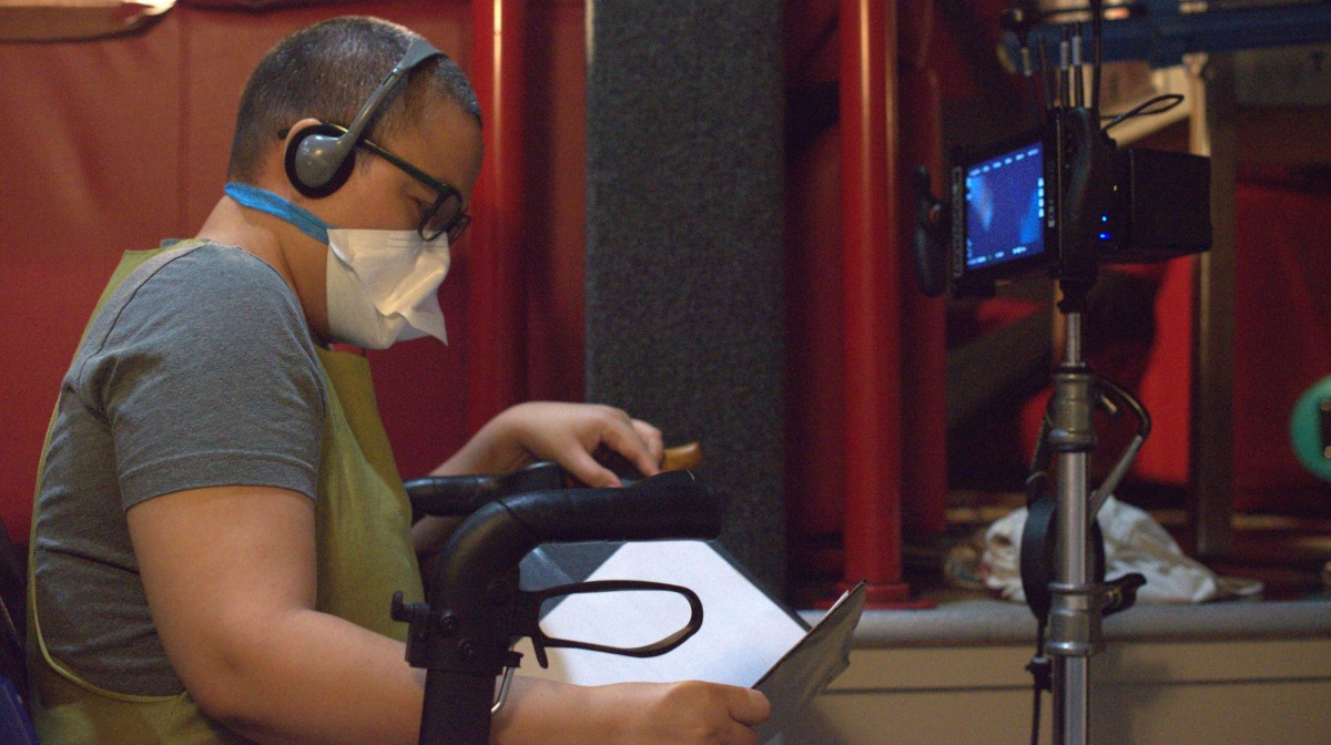 A person wearing headphones and a mask sits in a wheelchair, reading papers beside a mounted video camera.