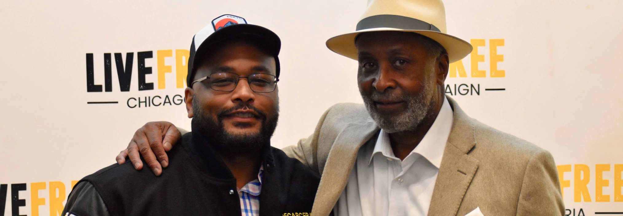 Two men posing together, smiling, in front of a 'LIVE FREE CHICAGO CAMPAIGN' step-and-repeat banner.