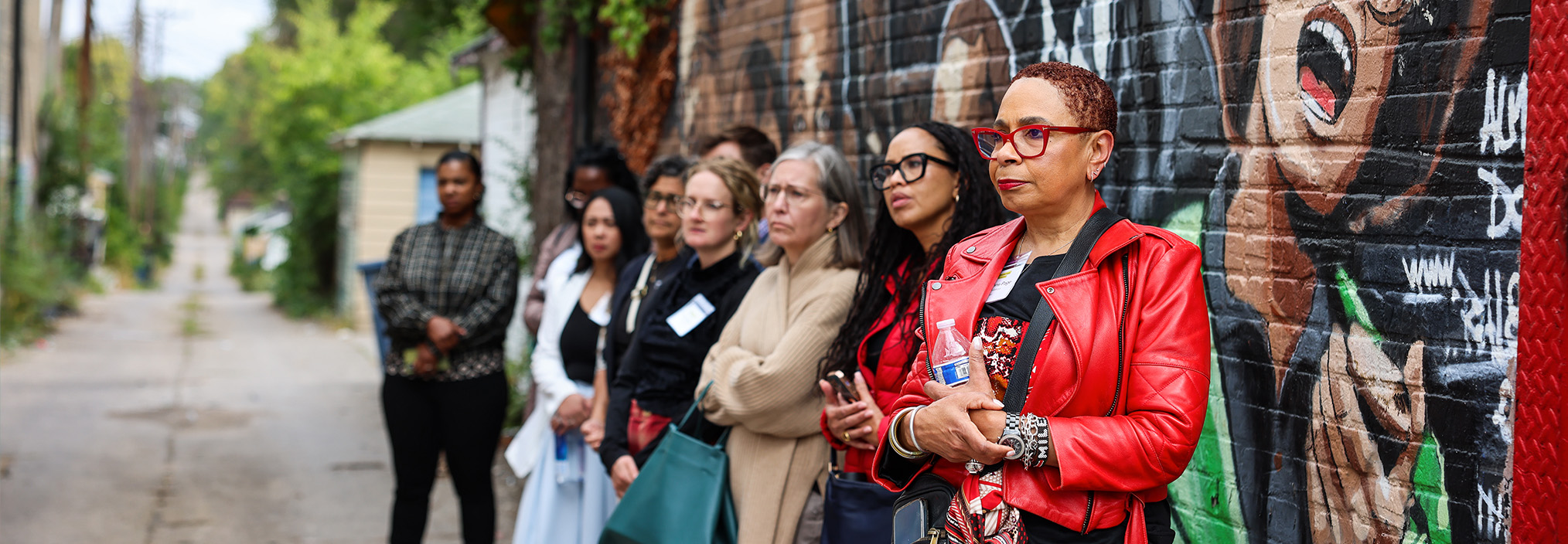 A group of women stand by a colorful mural on a brick wall in an urban alleyway, looking attentively ahead.